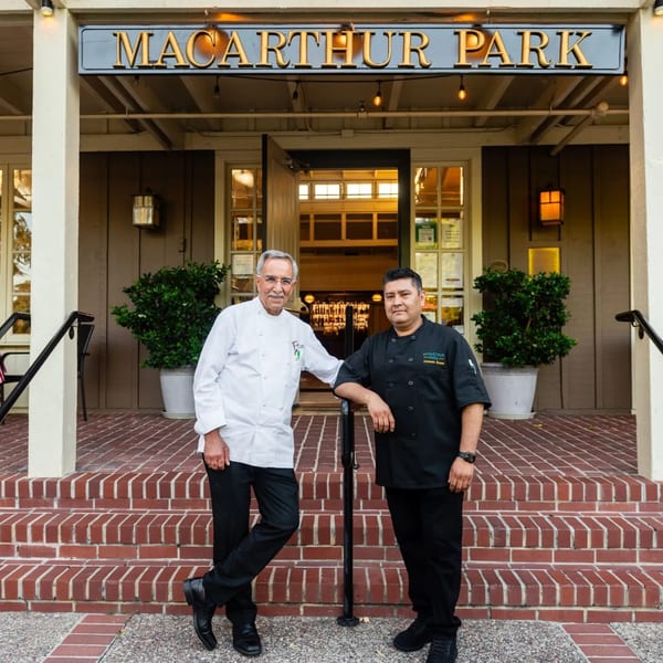 Two chefs, one older in white and one younger in black, stand on the brick steps of MacArthur Park Restaurant.