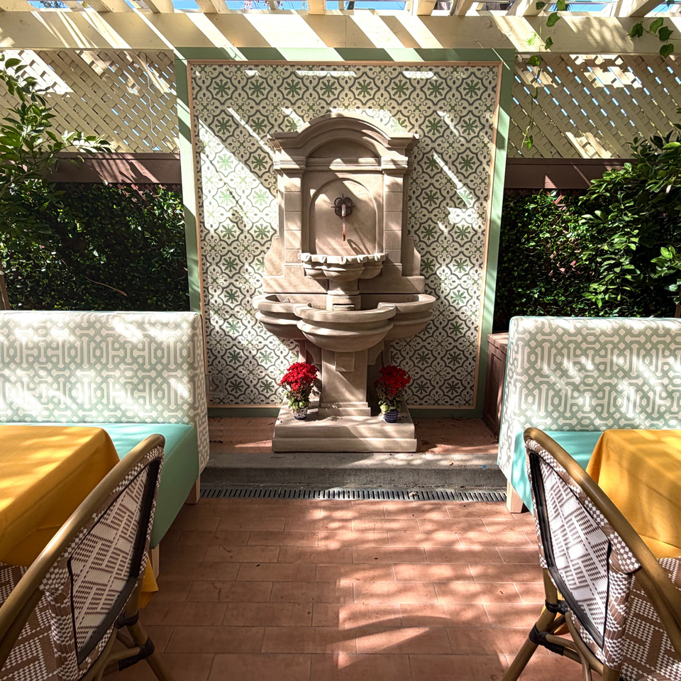 Sunlit outdoor restaurant patio with patterned tiled fountain, turquoise banquettes, yellow tables, greenery, and decorative latticework.