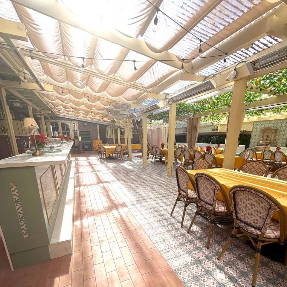 Sunlit restaurant patio with yellow tablecloths, patterned tile flooring, wicker chairs, greenery, and a decorative wall fountain.