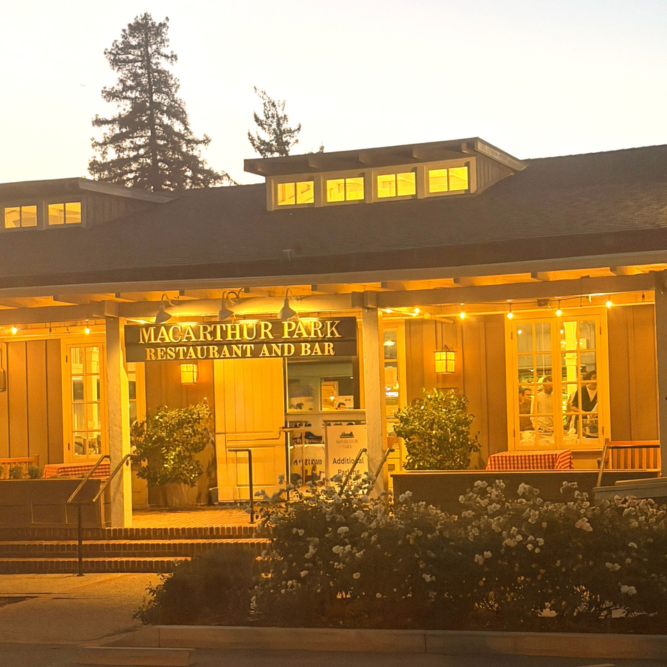 MacArthur Park Restaurant and Bar exterior glowing at dusk, with outdoor seating, string lights, flower beds, and tall trees.