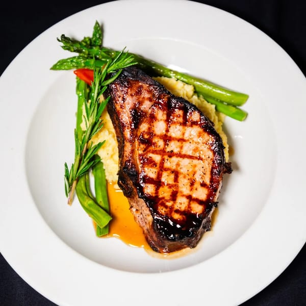 Grilled pork chop with mashed potatoes, asparagus, and a sprig of rosemary on a white plate.