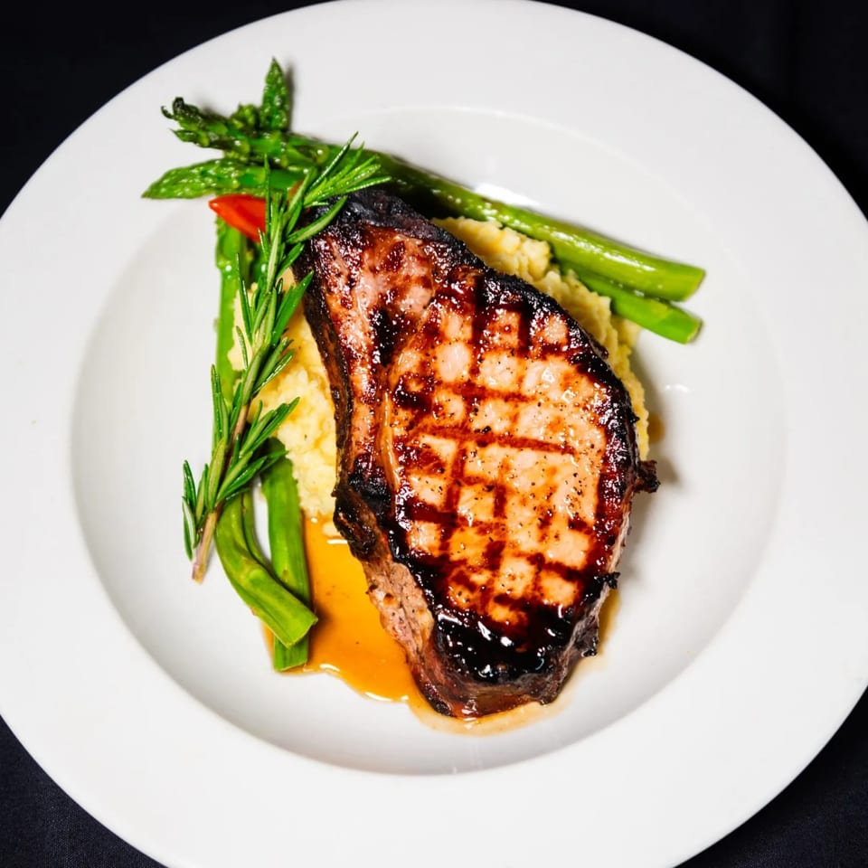 Grilled pork chop with mashed potatoes, asparagus, and a sprig of rosemary on a white plate.