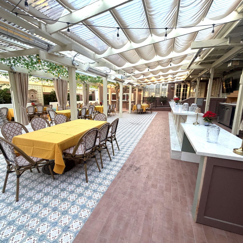 Bright restaurant patio with yellow tablecloths, patterned tile floors, draped canopy, string lights, and a marble service bar.