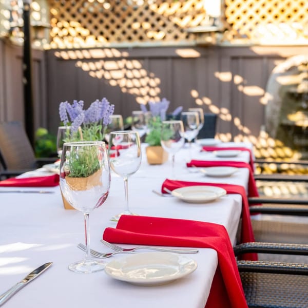 Outdoor dining table set with white tablecloths, red napkins, plates, and wine glasses, under a trellis.