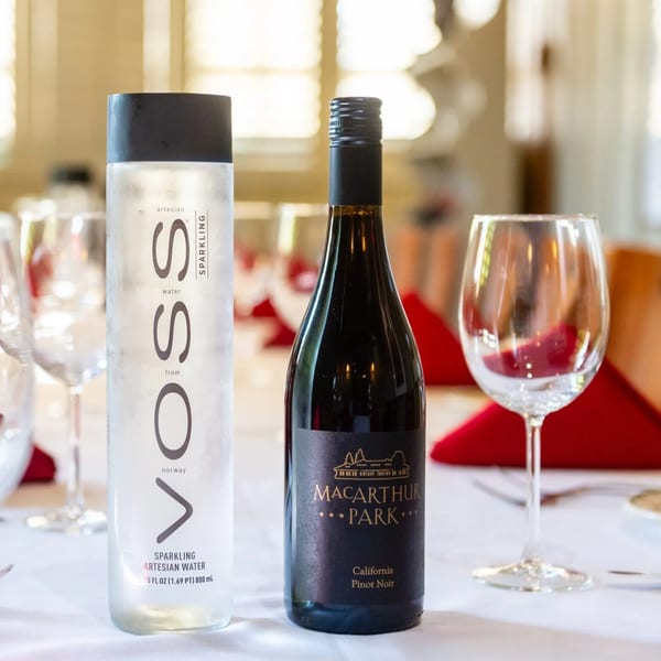 A bottle of MacArthur Park Pinot Noir and Voss sparkling water on a white tablecloth with wine glasses.
