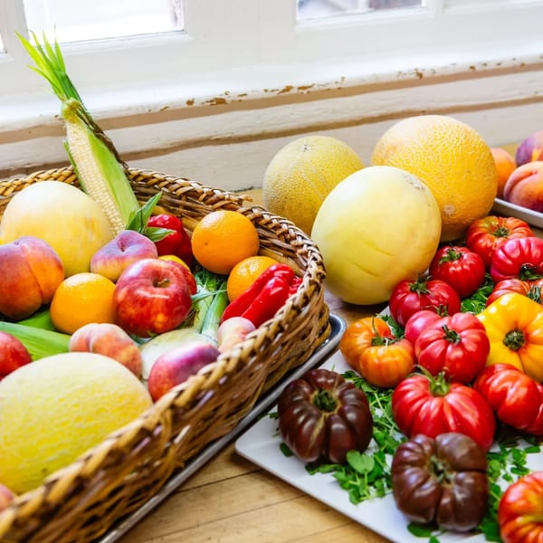 A wicker basket and white trays overflow with fresh peaches, melons, corn, tomatoes, and peppers.