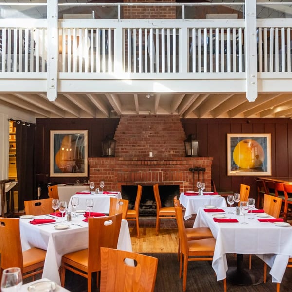 The MacArthur Park Restaurant dining room features set tables with red napkins, a brick fireplace, and a white balcony.