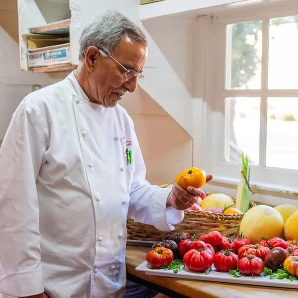 A chef in a white coat inspects a yellow tomato, with other colorful tomatoes and melons nearby.