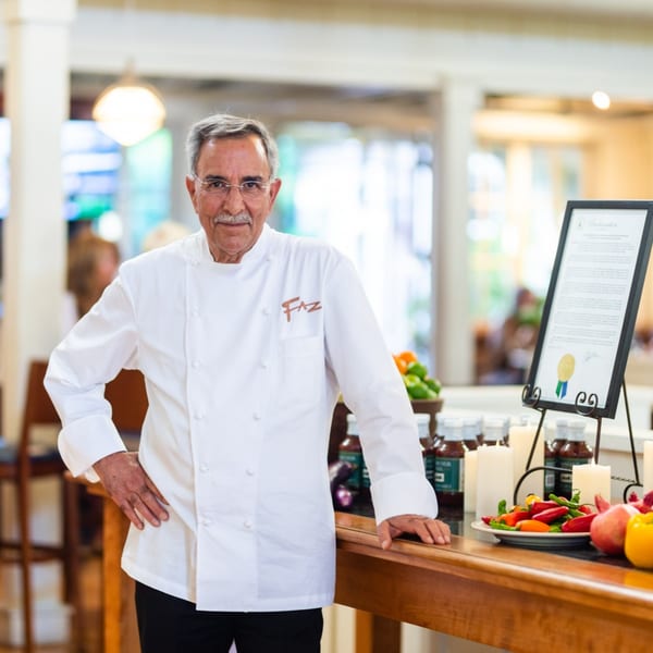 Chef Faz stands proudly in his white chef's coat next to a display of fresh produce and sauces.