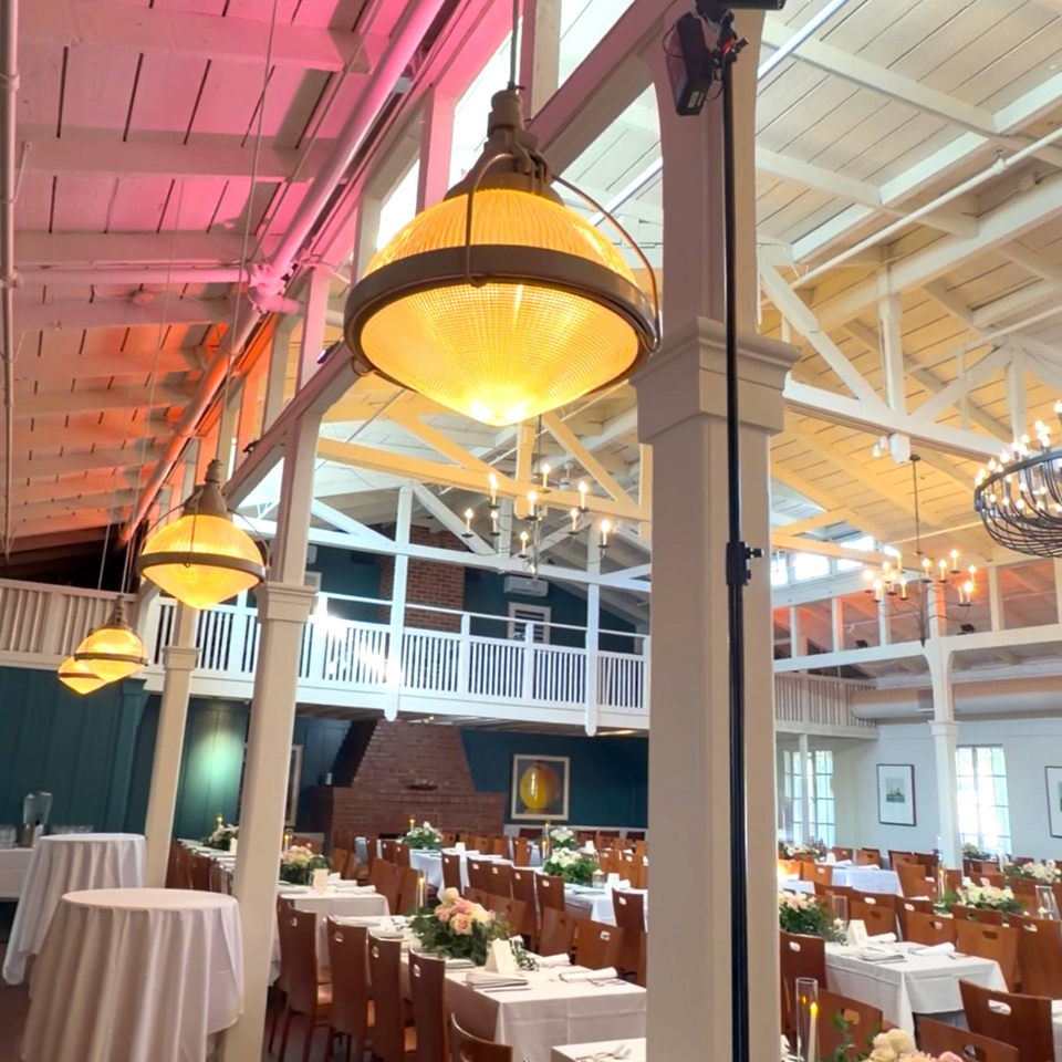 Elegant restaurant dining room with white tablecloths, floral centerpieces, exposed beams, chandeliers, and glowing pendant lights.