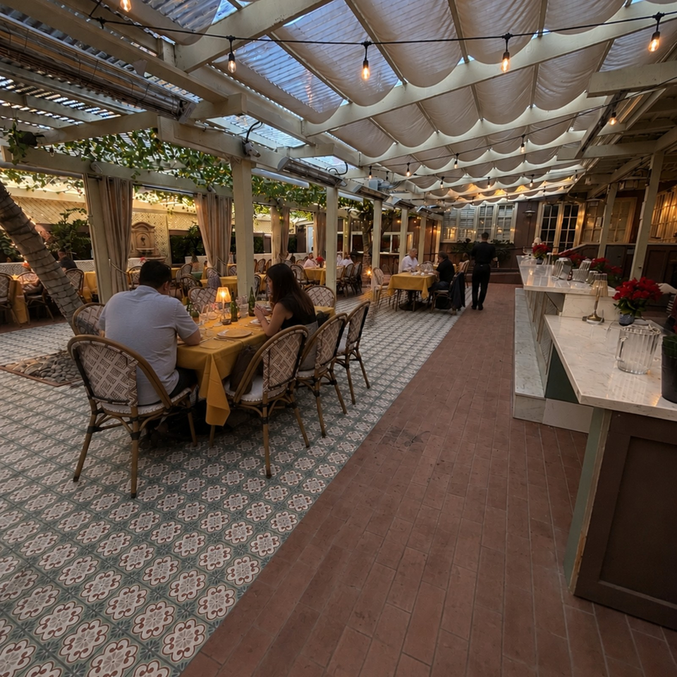 Elegant outdoor restaurant patio with patterned tile floors, diners, yellow tablecloths, tropical plants, and string lights beneath a canopy.