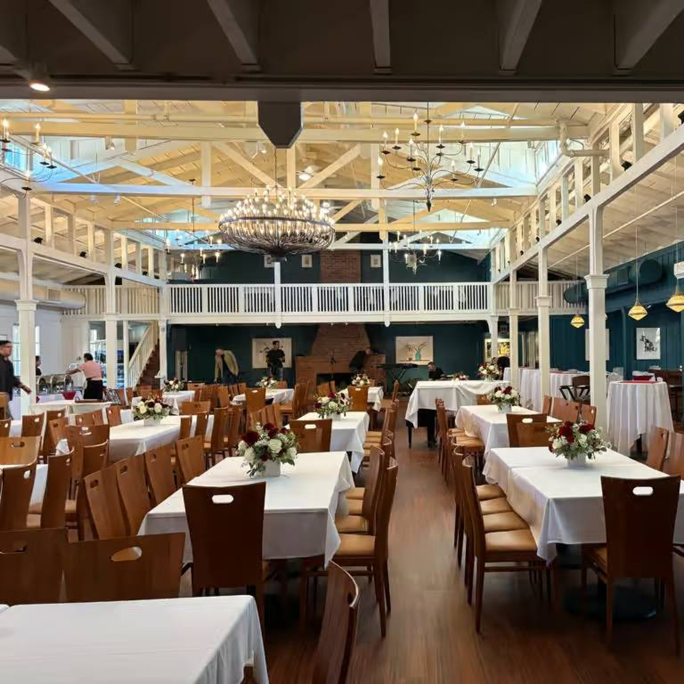 Elegant restaurant dining room with white tablecloths, floral centerpieces, wooden chairs, chandeliers, and a mezzanine balcony.