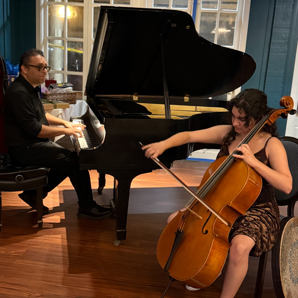 Pianist and cellist perform together indoors beside tall windows and polished wooden floors.