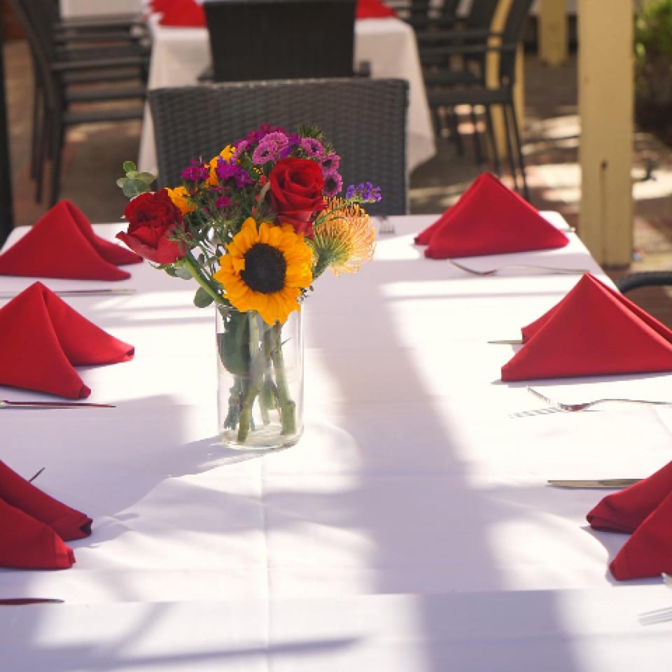Outdoor dining table set with white tablecloths, red napkins, and a colorful floral centerpiece.