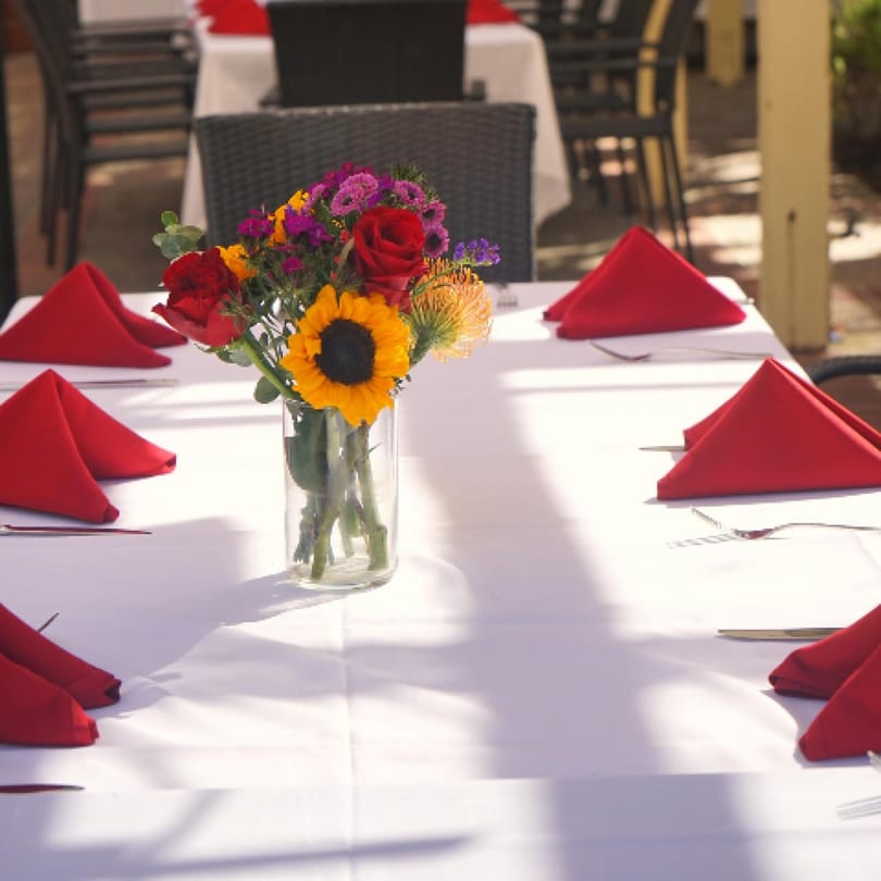 Outdoor dining table set with white tablecloths, red napkins, and a colorful floral centerpiece.